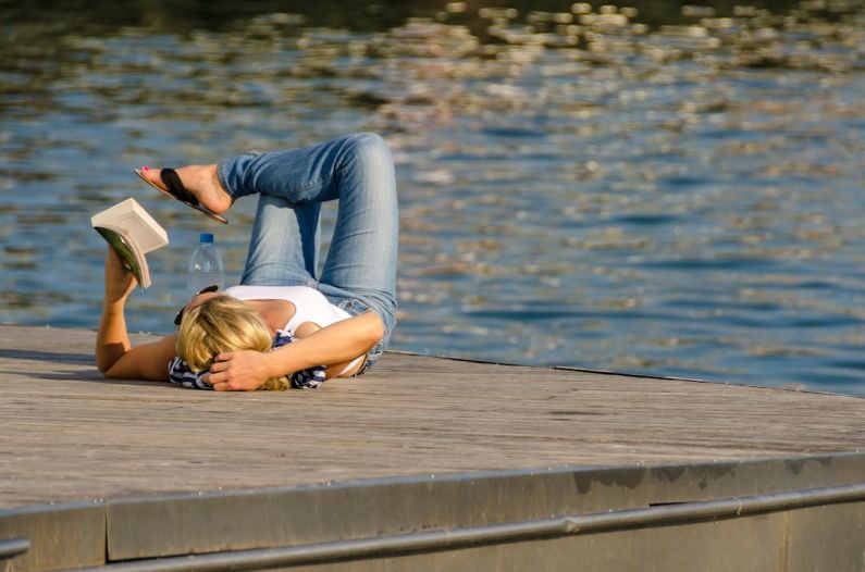 Downtime - woman in white tank top and gray pants lying on brown wooden dock during daytime